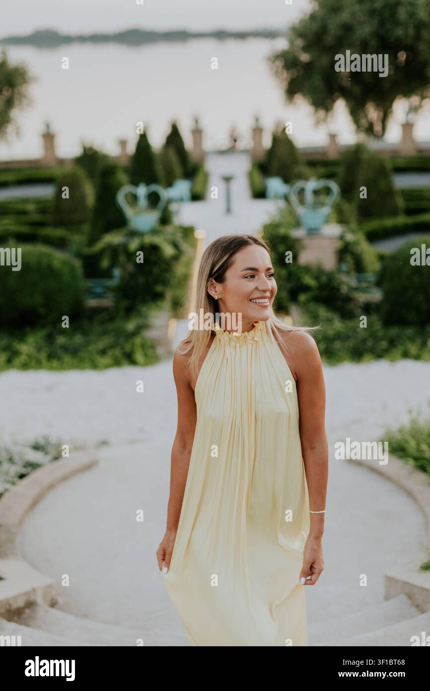 A woman walks down stone steps in a garden filled with plants. She smiles and wears a light dress. The sun sets behind the water, creating a soft glow Stock Photo