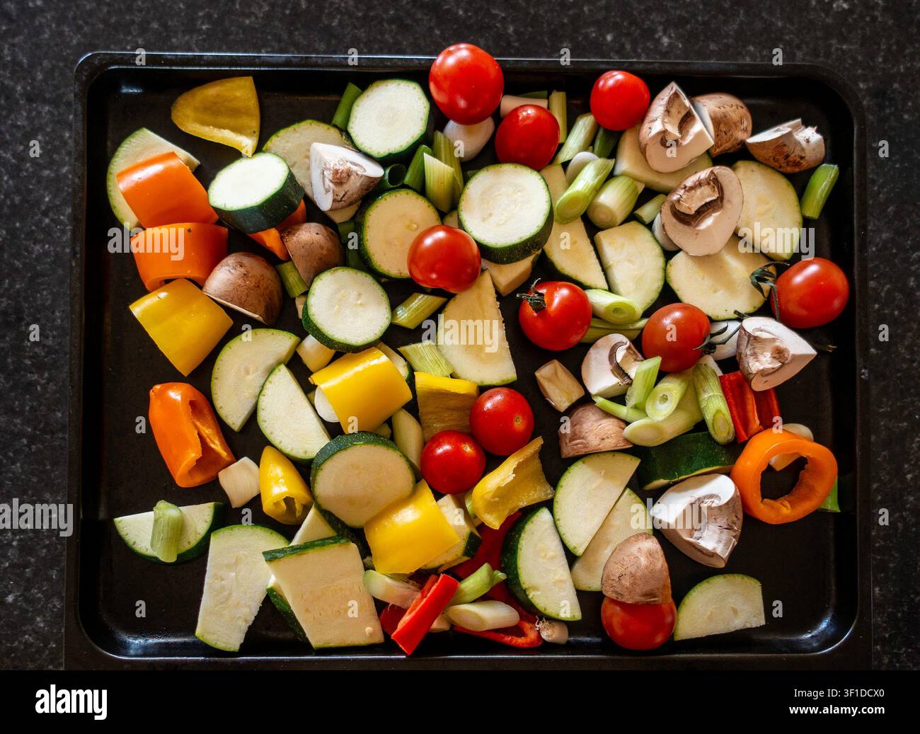 A variety of freshly chopped raw vegetables, including zucchini, cherry tomatoes, bell peppers, and mushrooms, arranged on a black baking tray. Stock Photo