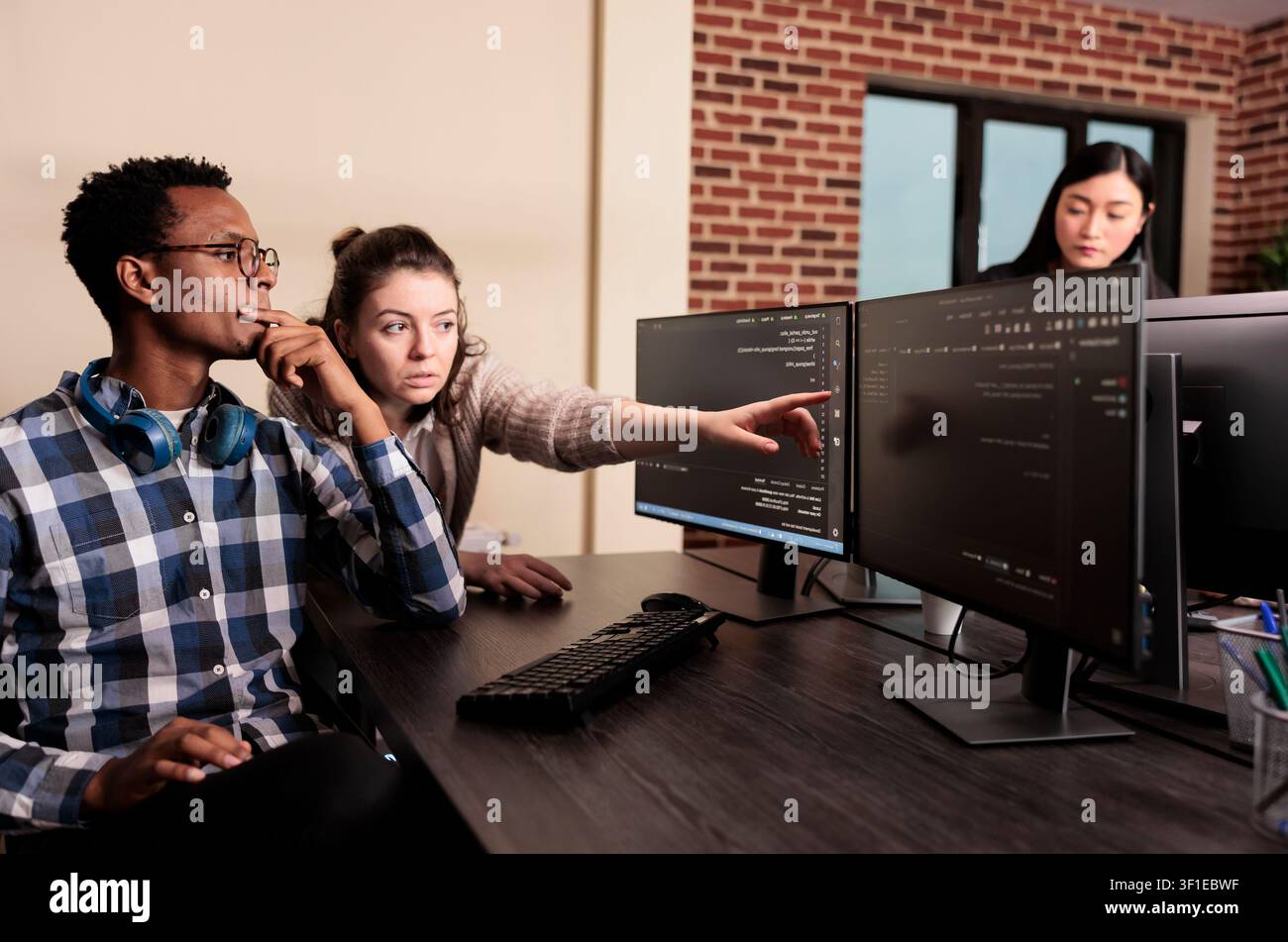 Software developer points to database script on a terminal window while asking a coder for help with HTML code in a data room, with developer teamwork, source code review. Stock Photo