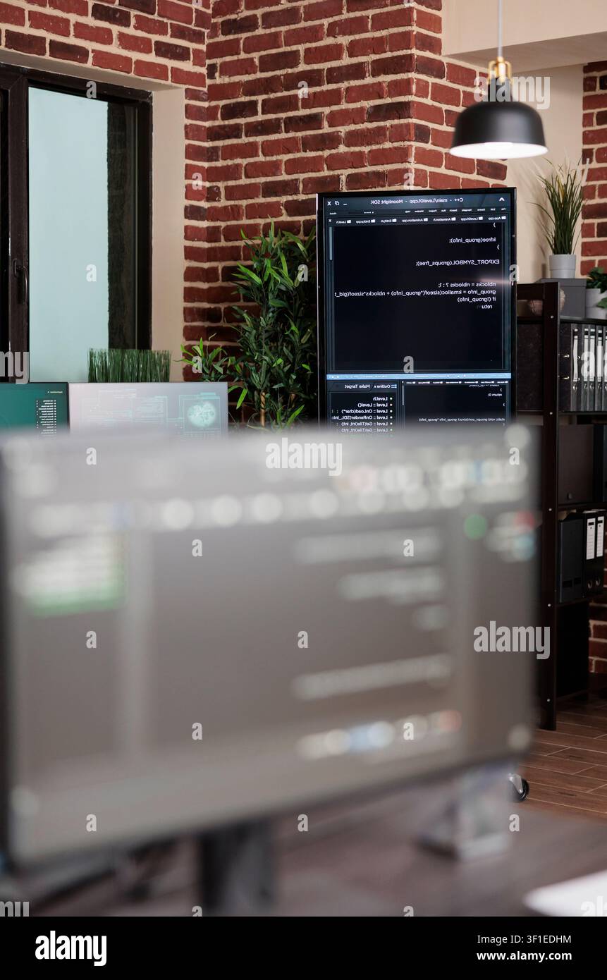 Empty data center office contains multiple computer screens showing programming code, server equipment and modern brick wall technology workspace. Stock Photo