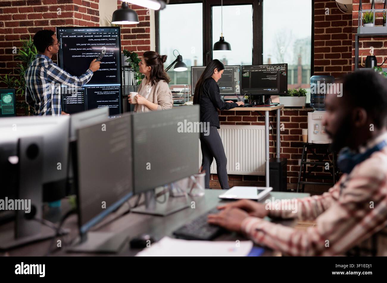 Technology team leader points out a source code mistake while software developers discuss an interface bug and slow data processing near a vertical screen. Stock Photo