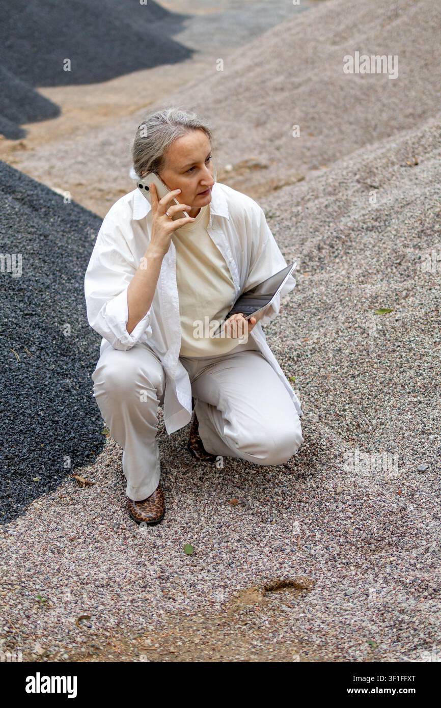Middle aged woman talks on smartphone while inspecting a construction aggregate stockpile and holding a tablet outdoors. Concept of field communicatio Stock Photo