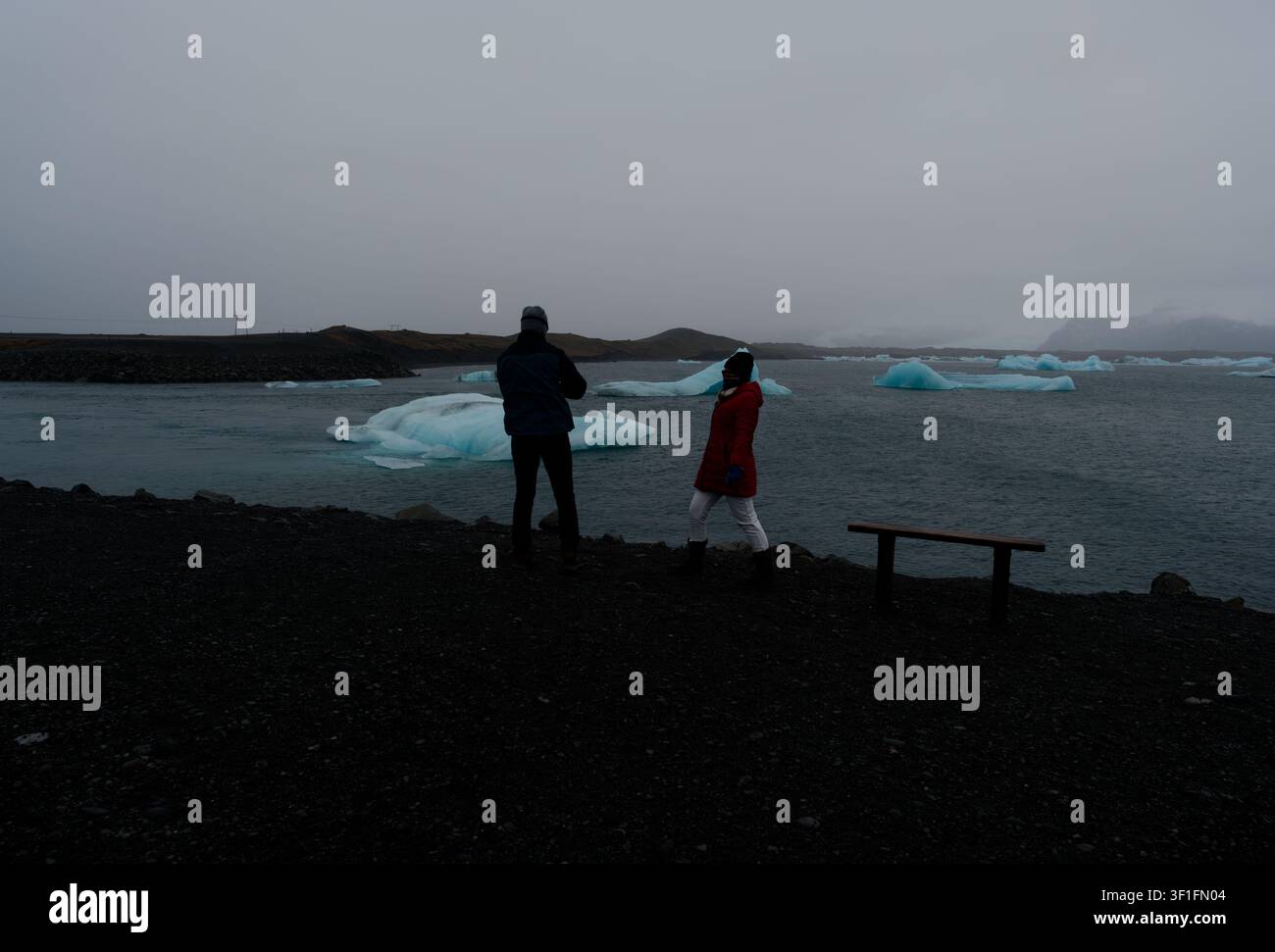 Two visitors walk the shoreline path of Jökulsárlón glacier lagoon with blue icebergs Stock Photo