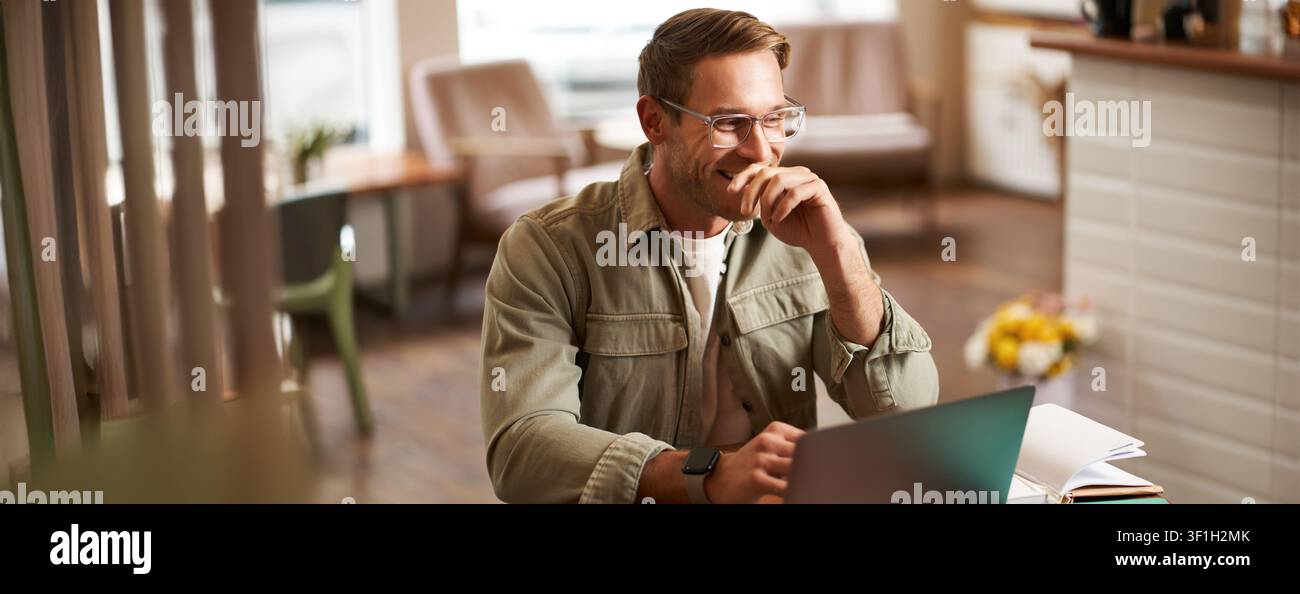 Image of young digital nomad, man in glasses sits in cafe, works from coffee shop, uses laptop in co-working space, wears glasses, drinks his beverage Stock Photo