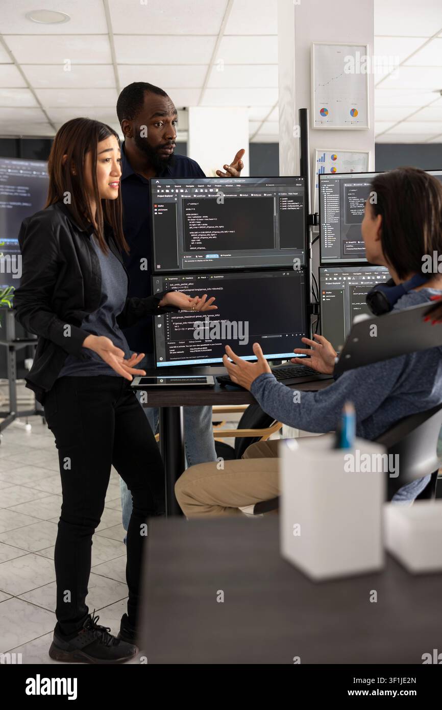 Diverse developers discuss programming data and source code on desktop pc during team meeting. Colleagues collaborate closely, brainstorming system improvements and digital problem solving strategies. Stock Photo