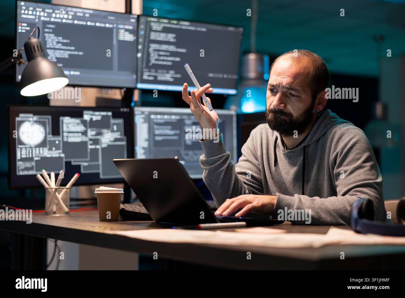 Developer stares at laptop with confused expression while holding pen and working on programming data. Multiple monitors display complex code, emphasizing challenges of technical decision making. Stock Photo