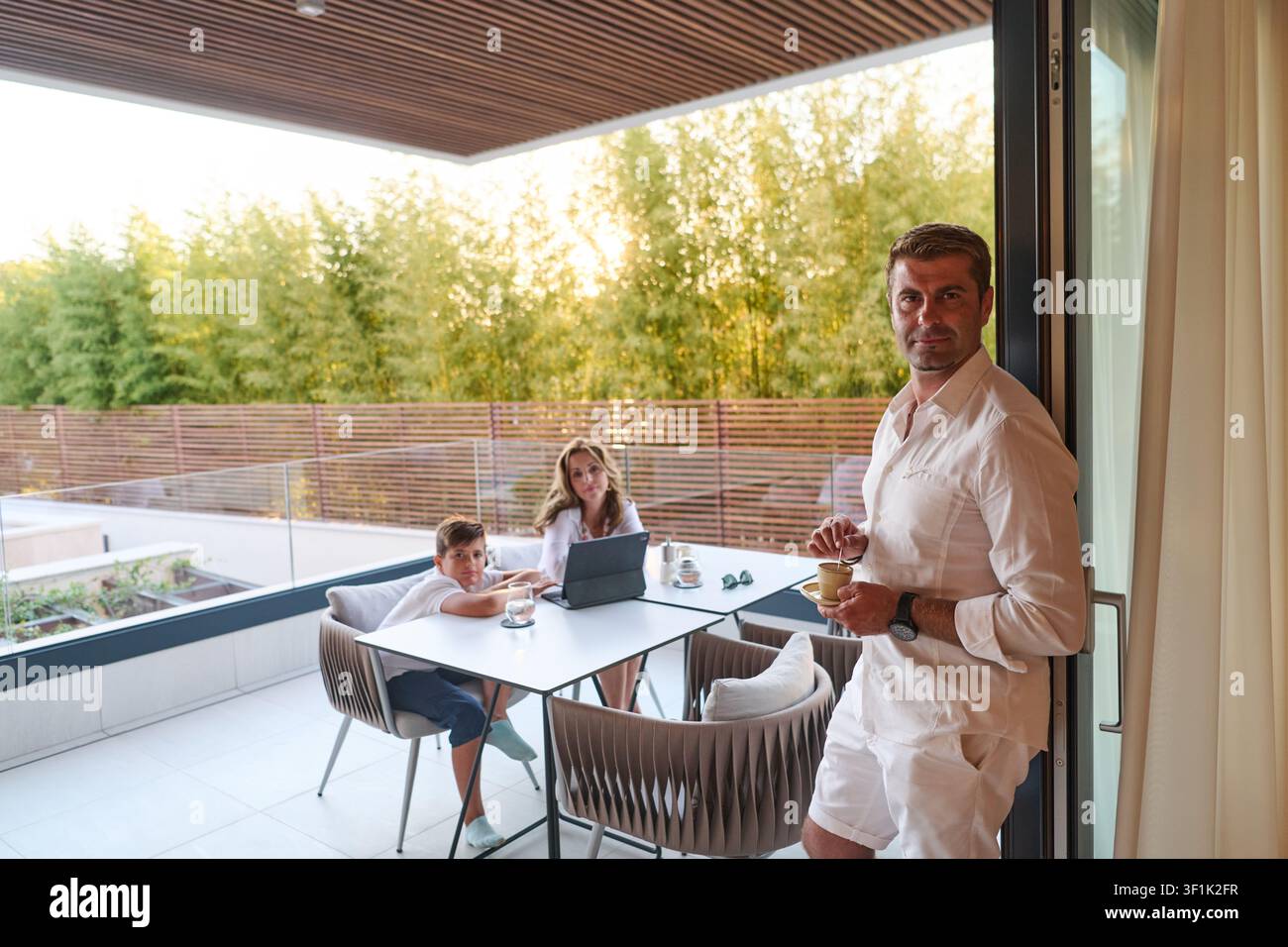 Family On Modern Balcony Terrace Having Coffee And Using Laptop While Father Stands At Door Stock Photo