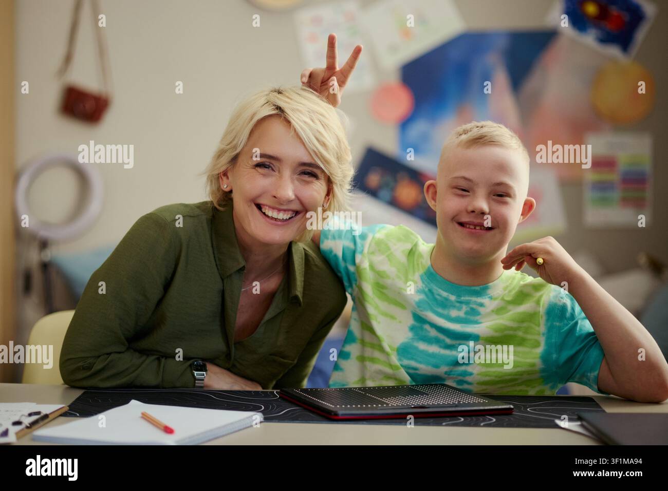 Portrait of Caucasian teacher and child with Down Syndrome smiling at camera, sitting at desk during classroom activity. Useful for inclusive education, support service, caregiving Stock Photo