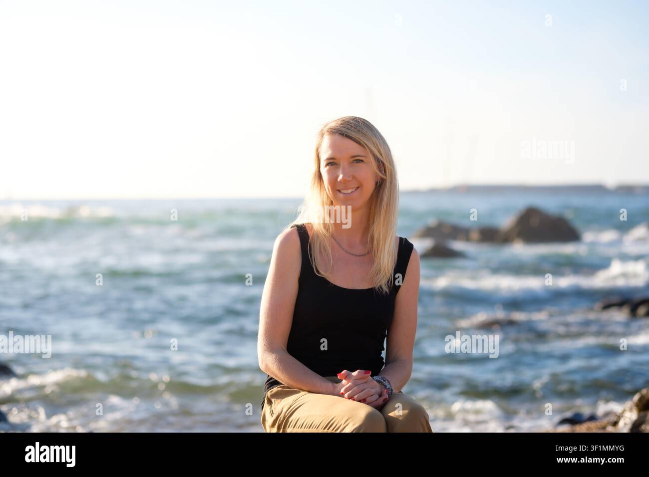 Woman sitting on rocks, enjoying sunlight and looking happily at the camera Stock Photo