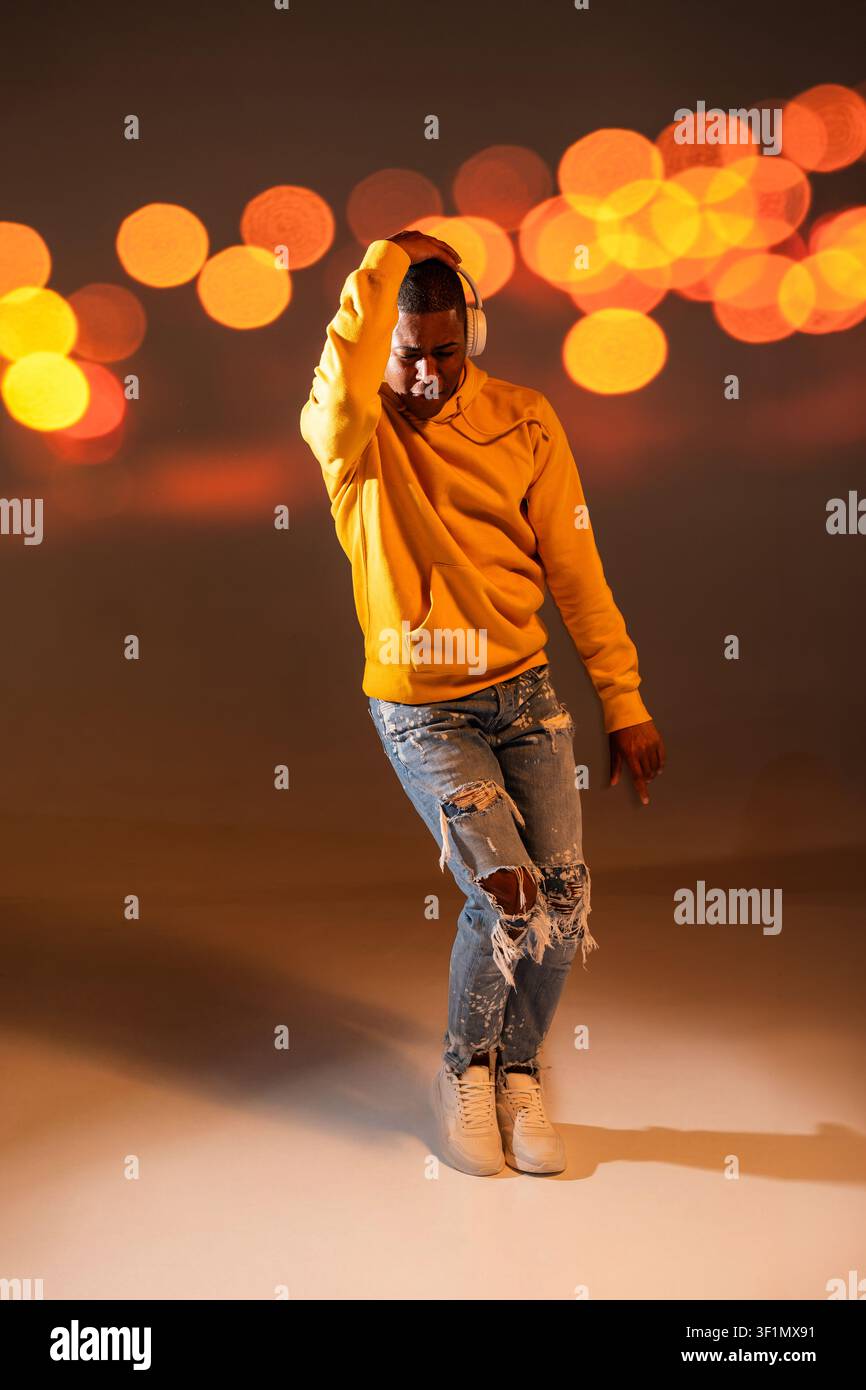 Full-length studio photo of a black man urban dancer wearing a yellow hoodie and ripped jeans, posing with headphones against warm bokeh lights, moder Stock Photo