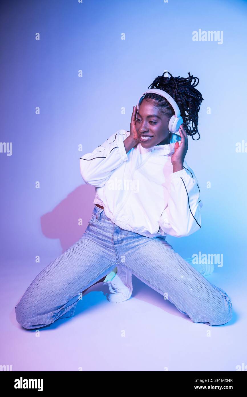 Afro woman dancer kneels in a modern studio, smiling while holding headphones and moving to the beat. Colorful blue and pink lighting creates a vibran Stock Photo