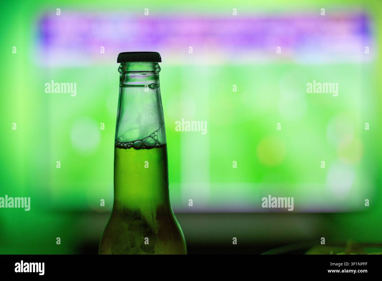 Close-up of a cold beer bottle with a blurred TV screen showing a live football match in the background. Vibrant green ambient backlight. Perfect for Stock Photo