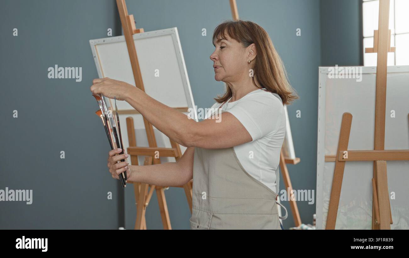 Woman painter selects paintbrushes and adjusts brush tips before blank canvas in studio; creative focus. Stock Photo
