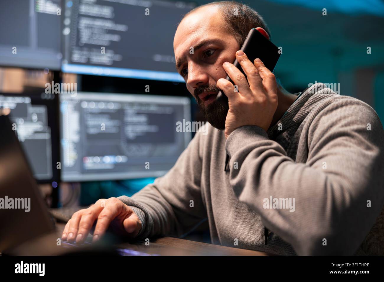 Cybersecurity specialist speaks on cellphone while typing at workstation, surrounded by multiple monitors displaying code. Programmer focused on addressing website issues and providing client support. Stock Photo