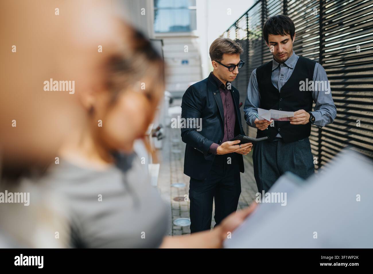 Two businessmen reviewing documents outdoors while a colleague reads notes in foreground Stock Photo