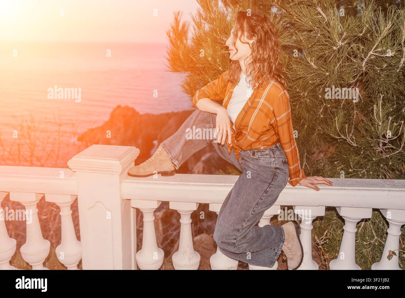 Woman balcony sunset sits on a white railing overlooking the ocean during golden hour for a peaceful vacation mood Stock Photo