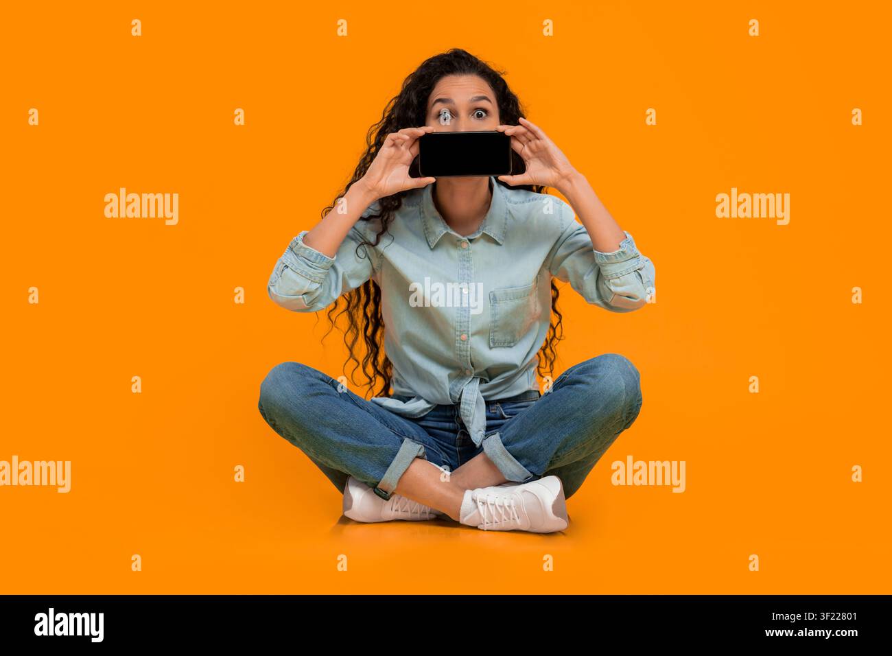 Young Woman Holds Smartphone in Front of Her Face While Sitting on Bright Background Stock Photo