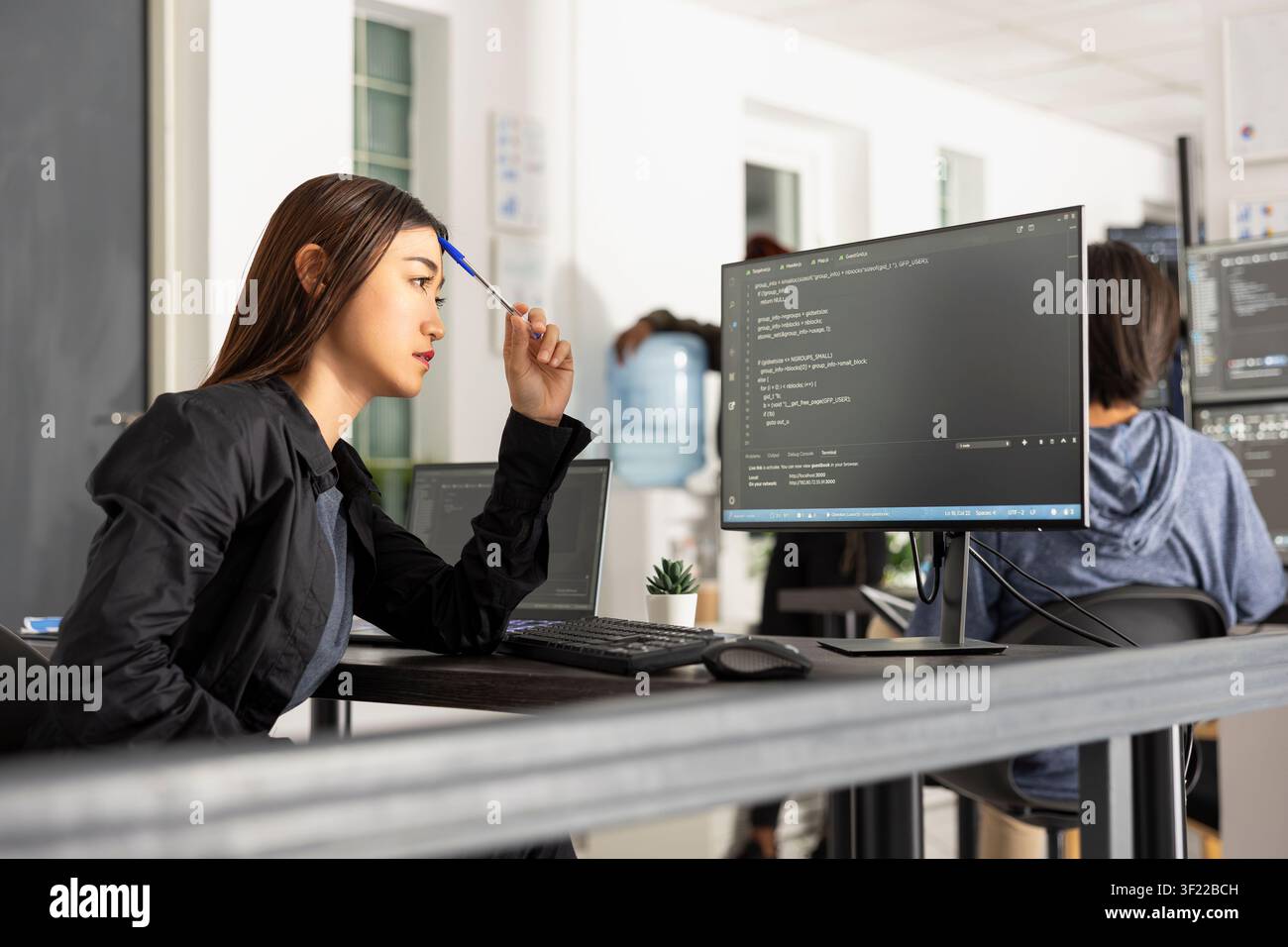 Female asian programmer works at desk, tapping pen while analyzing code on computer screen. Scene conveys concentration, innovation and digital workflow in modern software and web development. Stock Photo