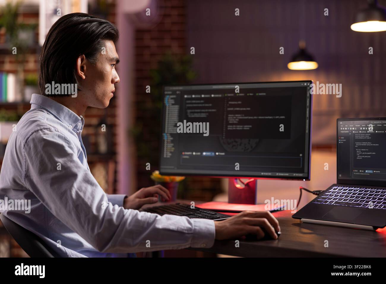 Young male entrepreneur spending late evening hours at home improving app performance and functionality. Focused programmer working from home and analyzing html code displayed across computer screens. Stock Photo