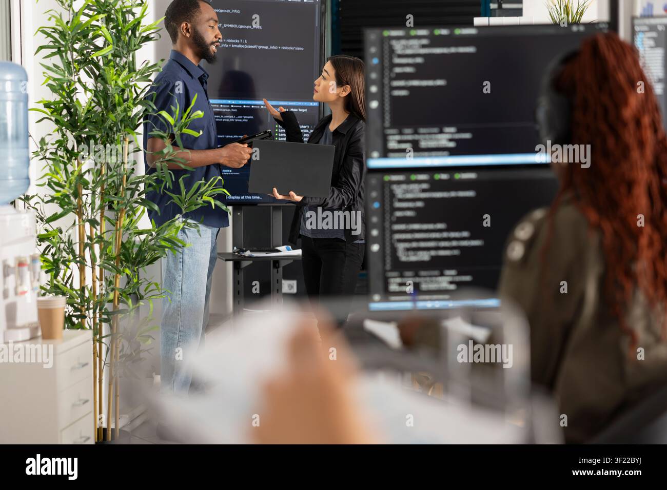 Male and female software developers stand by computer monitors, brainstorming coding project for startup. Diverse team collaborates on problem solving HTML scripts while building and refining website. Stock Photo