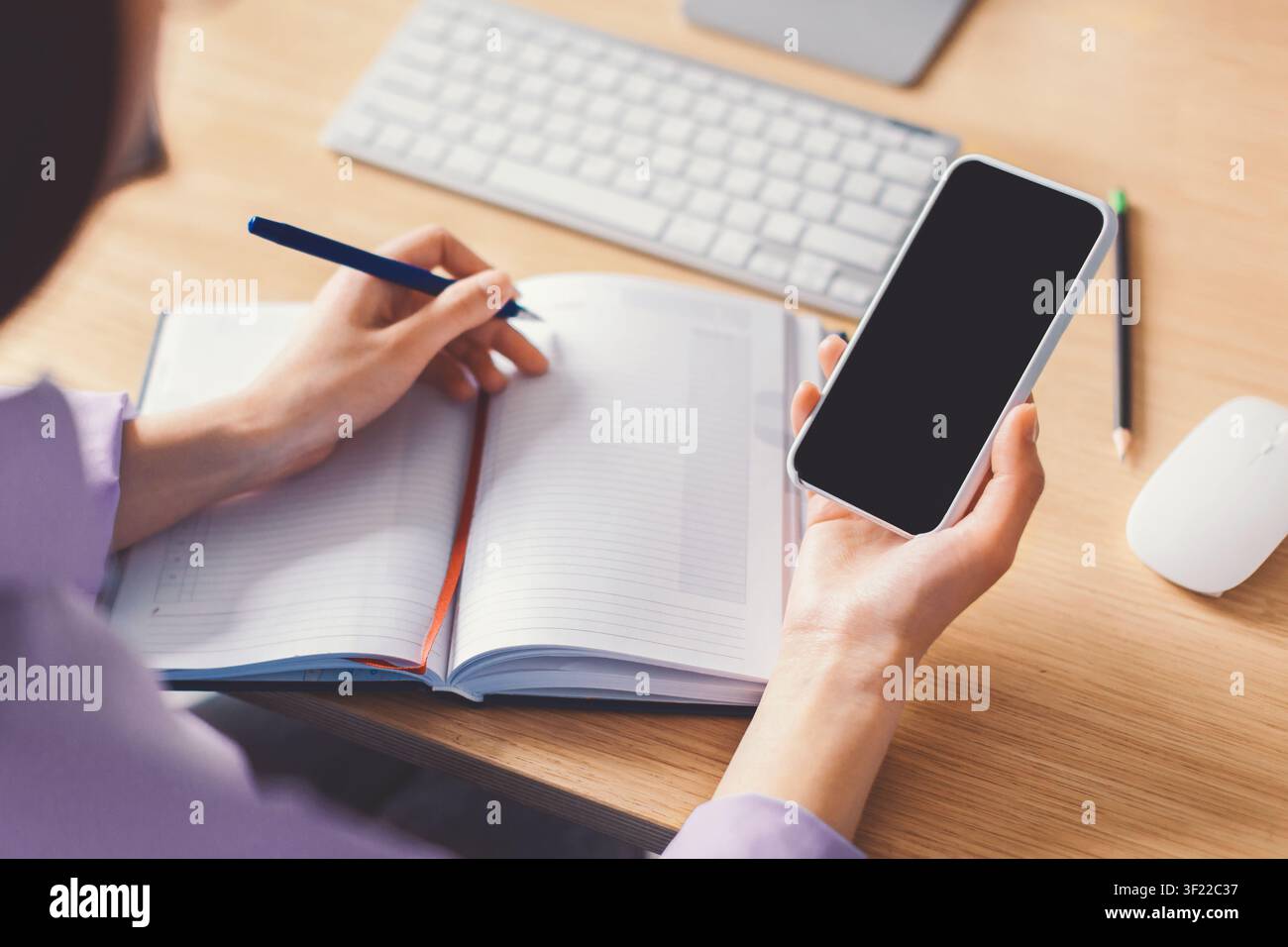 Unrecognizable Person Writing Notes In Journal While Holding Smartphone, Office Setting Stock Photo