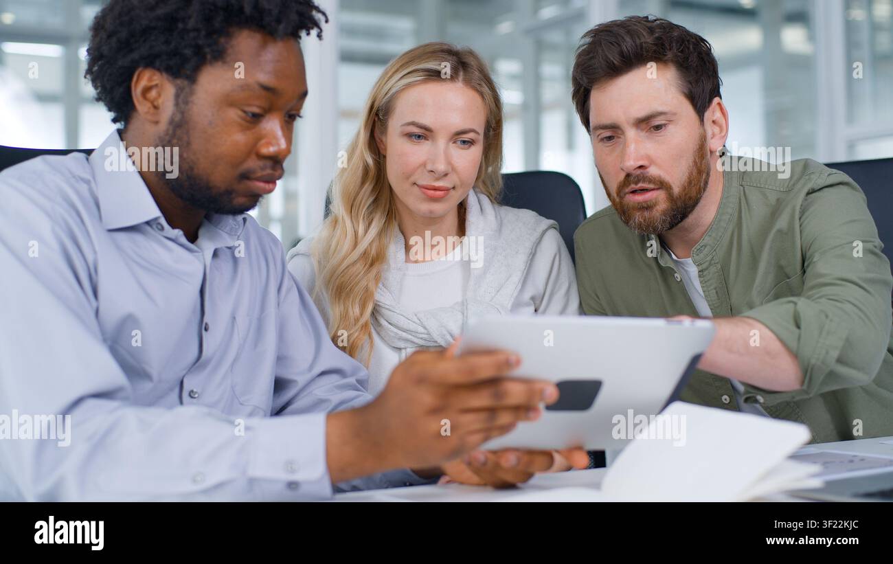 Happy multiethnic business team looking in digital tablet app smiling laughing talking three business colleagues using portable computer with Stock Photo