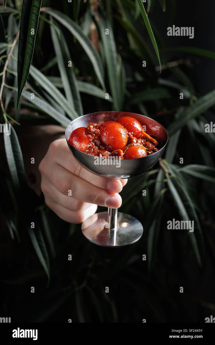 A close-up of a hand holding a stylish cocktail glass filled with pickled tomatoes, surrounded by lush green leaves, perfect for culinary and beverage Stock Photo