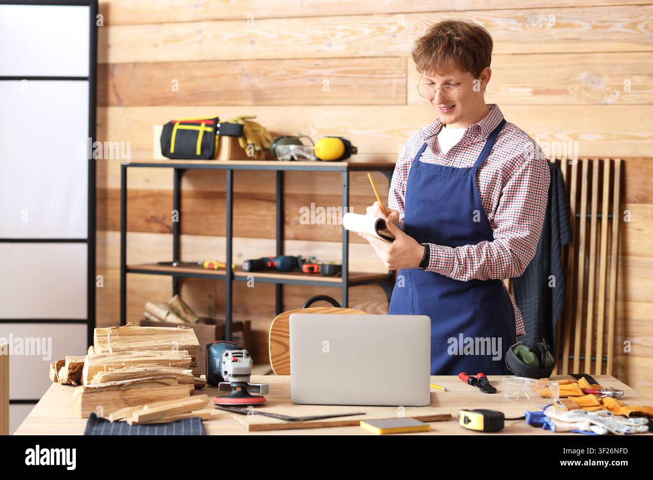Young carpenter with notebook using laptop at table in workshop Stock Photo