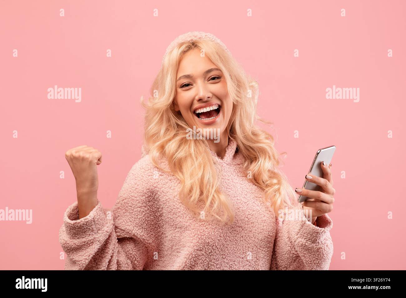 Happy Young Woman Celebrating With Smartphone In Hand, Joyful Expression, Pink Background Stock Photo