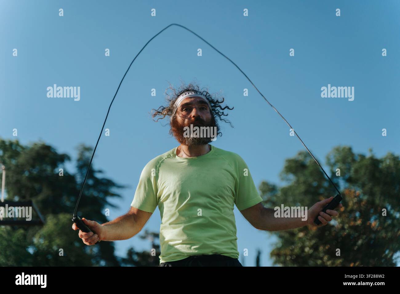man actively jumping rope in an outdoor setting on a sunny day, surrounded by trees, wearing a green t-shirt and black shorts, captured from a low angle that emphasizes his energetic motion. Stock Photo