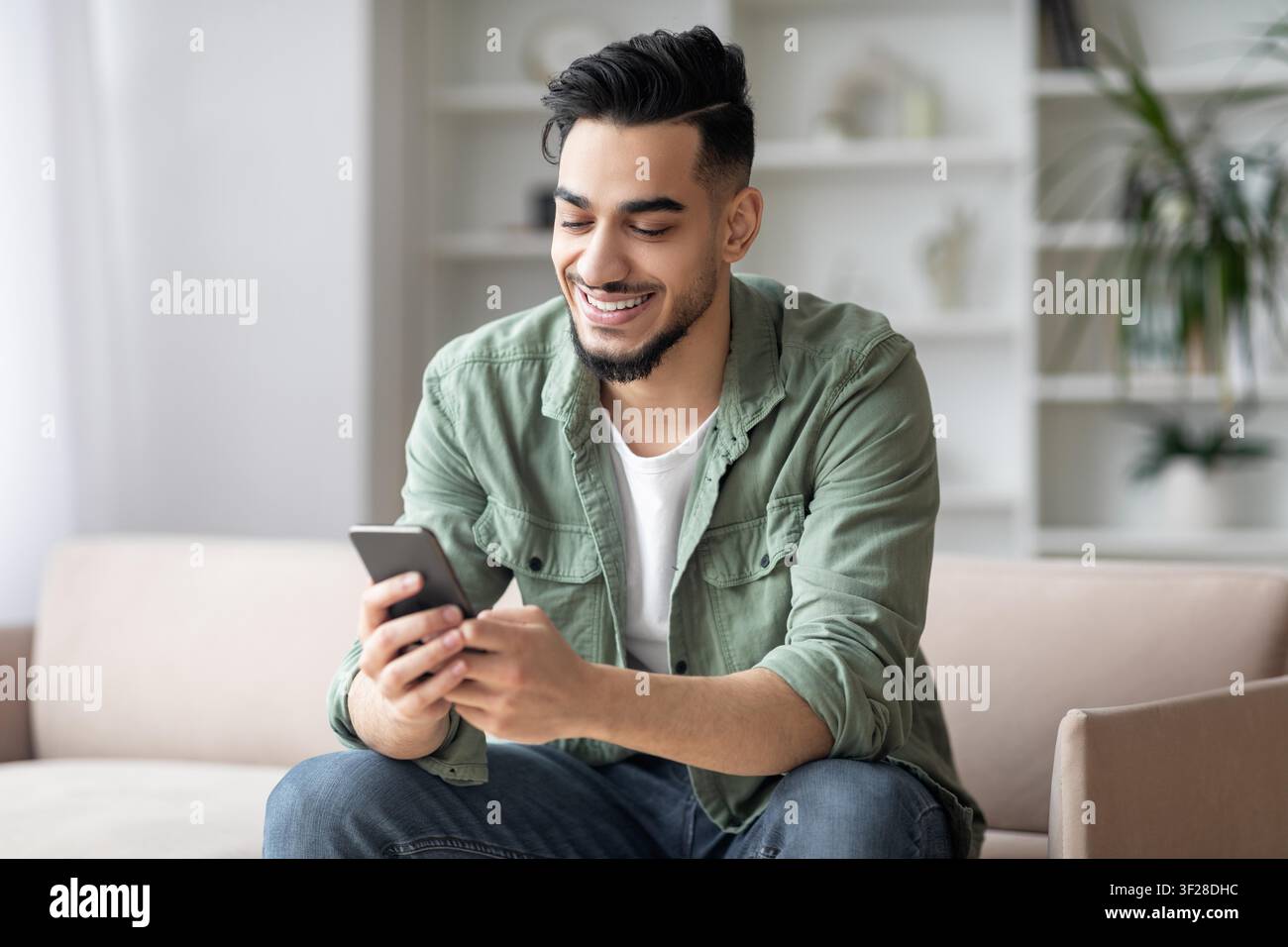 Smiling Young Asian Man Using Smartphone At Home, Relaxed Indoor Atmosphere Stock Photo