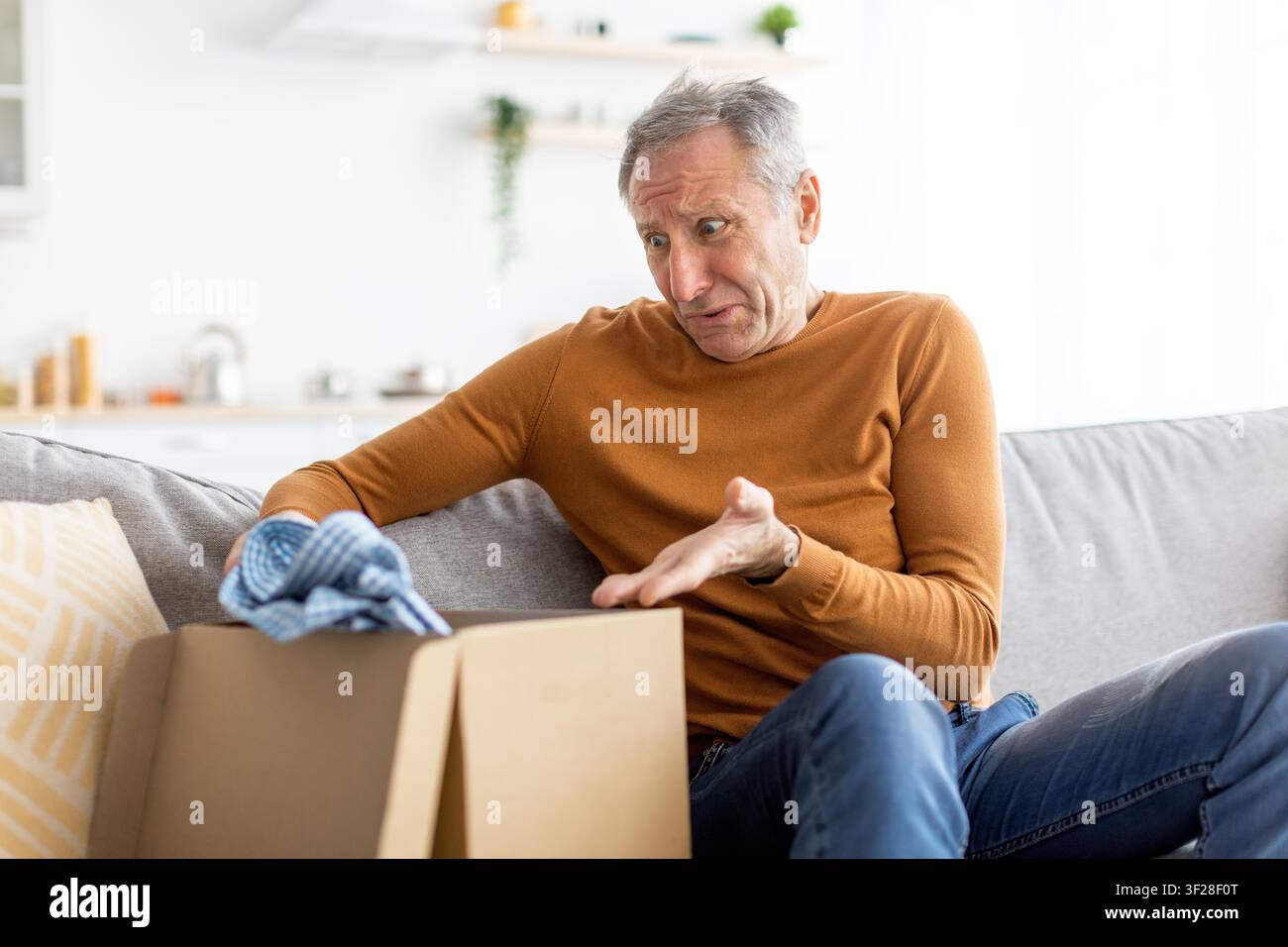 Worried Elderly Caucasian Man Sitting On Sofa, Opening Package At Home, Surprise Expression Stock Photo