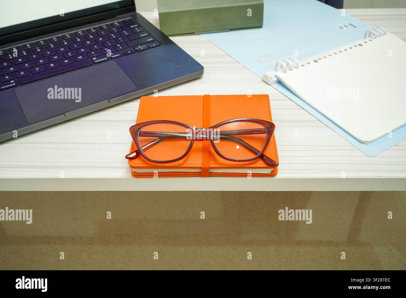 Stylish eyeglasses placed on orange notebook beside laptop and open notepad on white desk, ideal for study and remote work concepts. Stock Photo