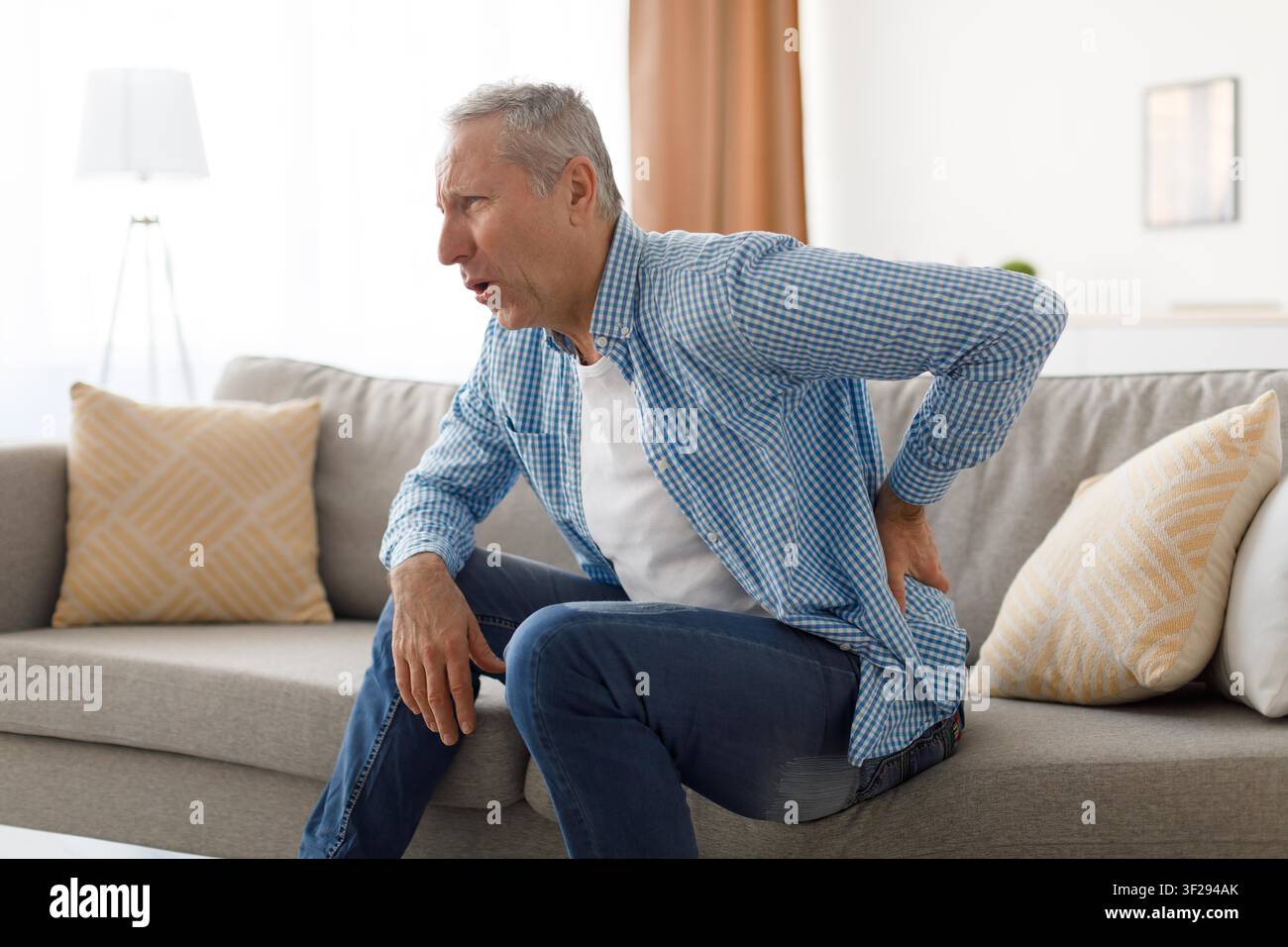 Elderly Caucasian Man Experiencing Back Pain While Sitting On Couch, Healthcare Concept Stock Photo