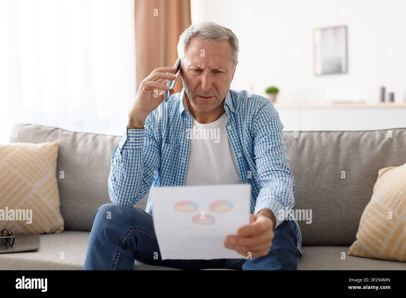 Focused Elderly Caucasian Man Analyzing Document While Talking On Phone, Home Office Stock Photo