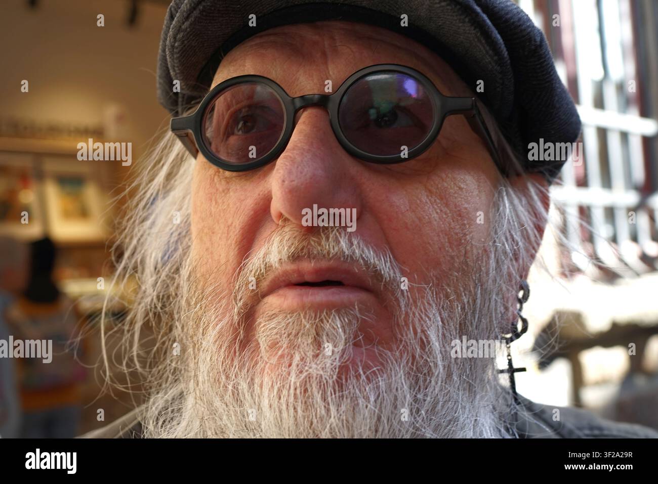 An senior man is shown with a beard, wearing round black glasses and a dark cap. His expression reflects varied emotions. A cross earring is visible o Stock Photo