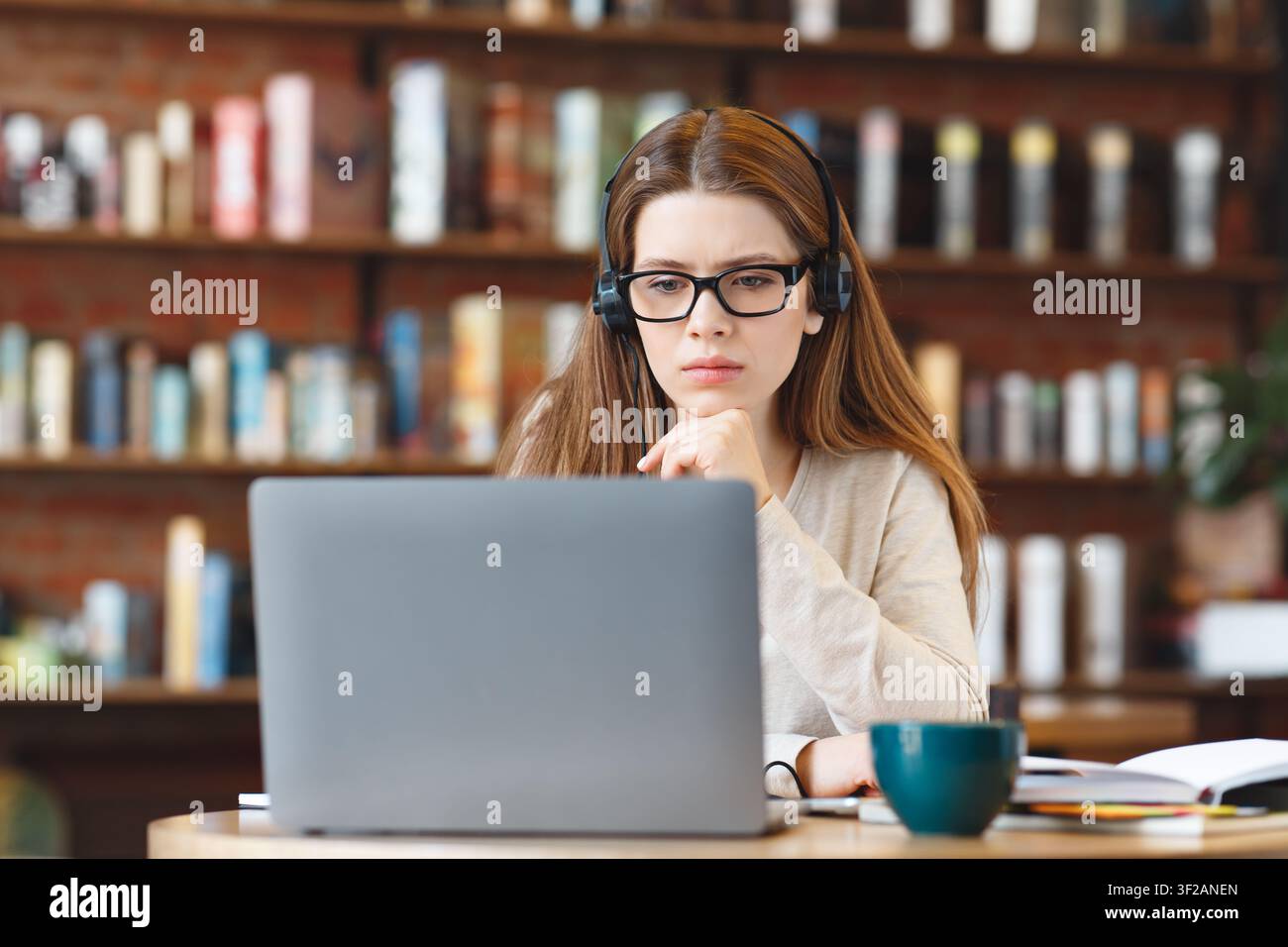 Focused Young White Woman Working On Laptop With Headphones In Modern Coffee Shop Setting Stock Photo