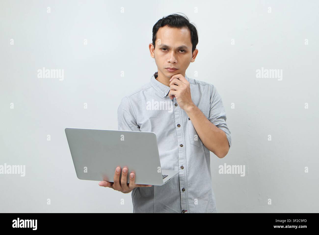 Pensive young Asian Indonesian man holding laptop computer with a serious face thinking about a question, thoughtful about confusing idea on isolated Stock Photo