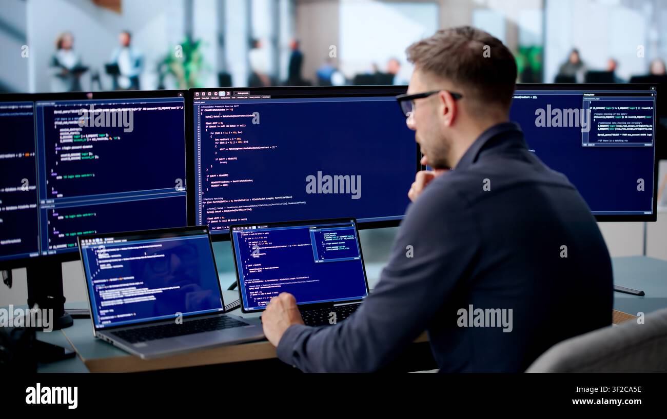 Young Software Engineer Coding Secure Algorithms On Laptop At Busy Office Desk. Stock Photo