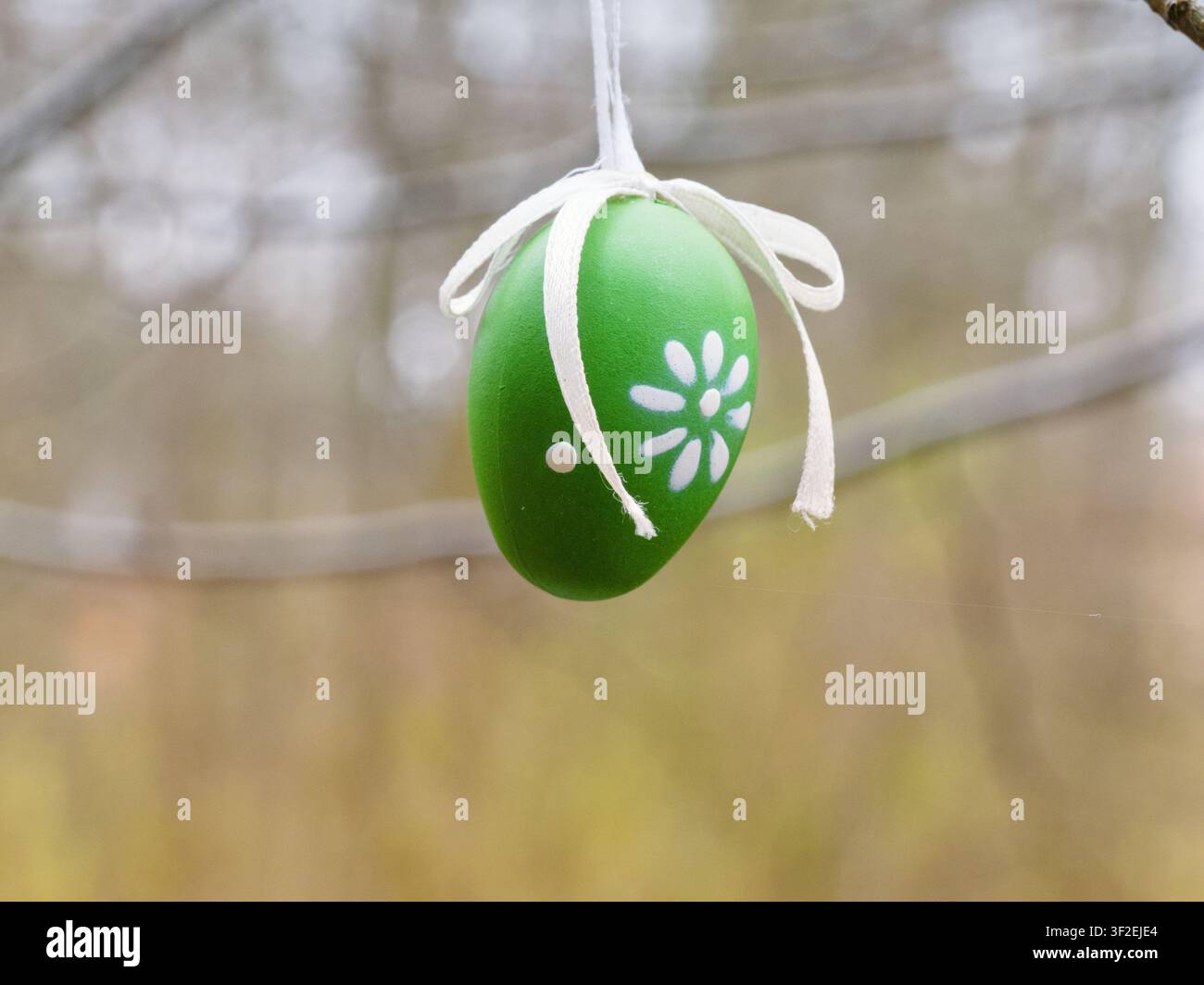 Decorative green painted Easter egg with white flower pattern hanging on a tree branch outdoors. Stock Photo