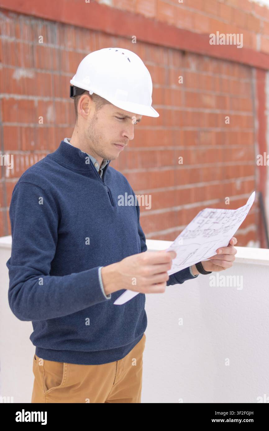 Construction professional wearing a safety hard hat, carefully examining architectural plans and blueprints on a building site, overseeing work progre Stock Photo