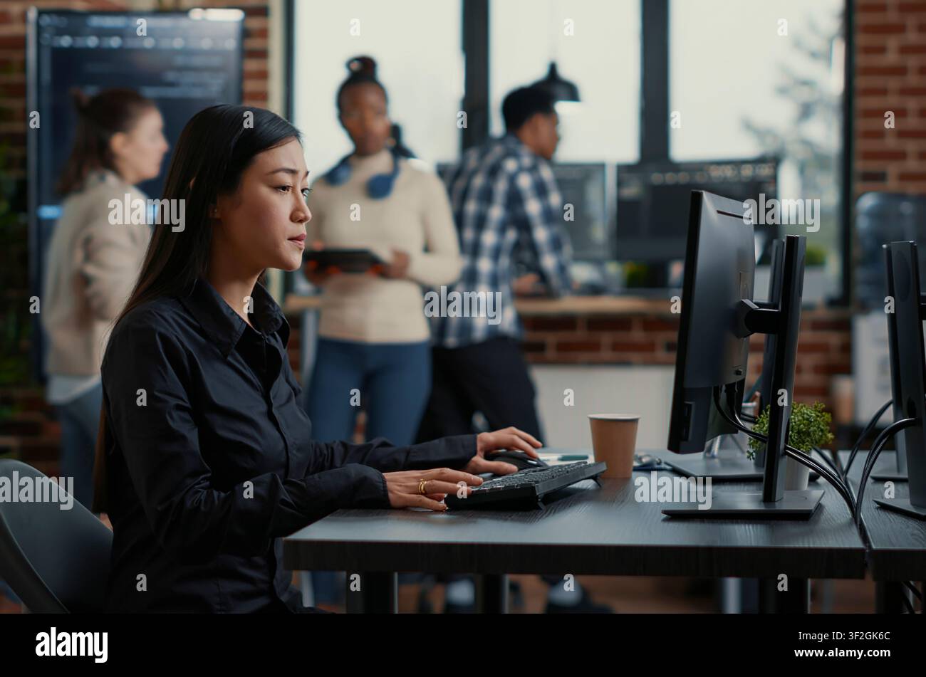 Asian software developer sits at a desk with a clipboard and starts typing on a computer keyboard while programmers analyze code on a wall screen. Stock Photo