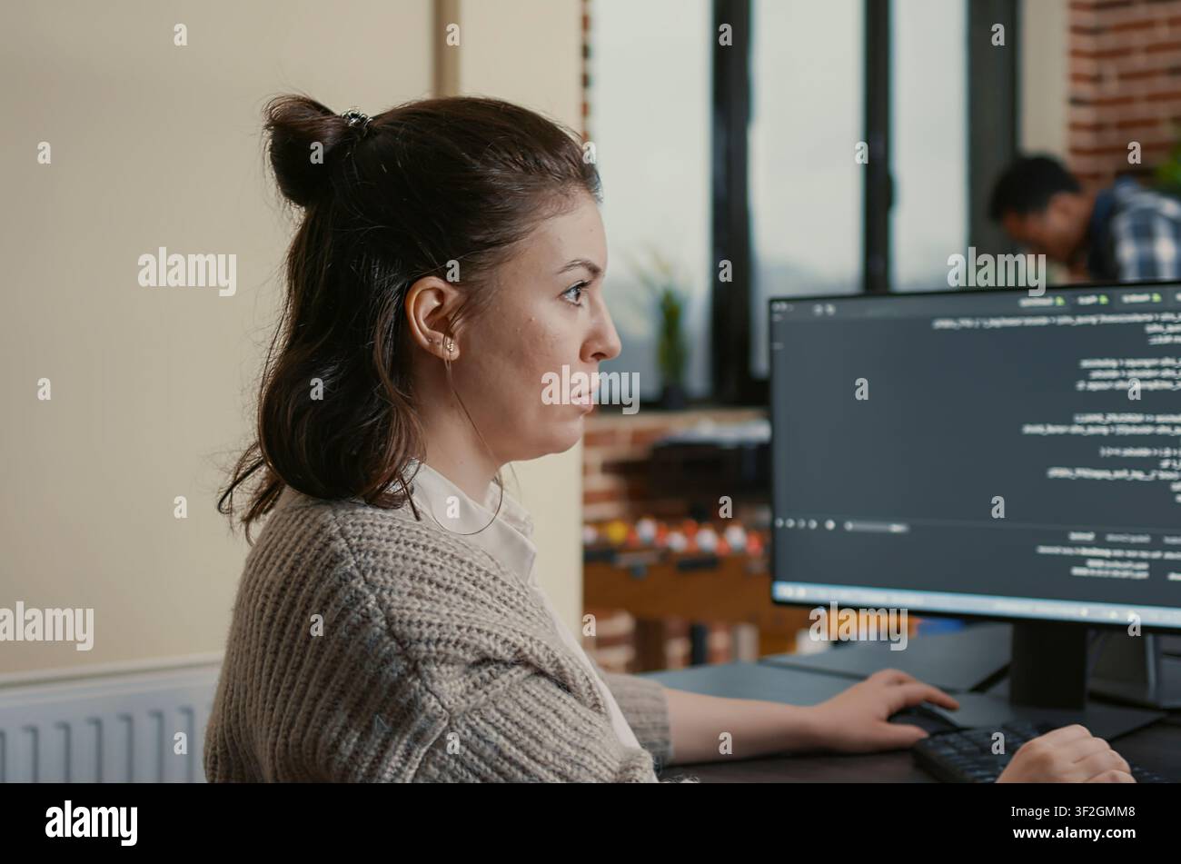 Focused software engineer writes code while viewing multiple computer screens with programming language algorithms as colleagues collaborate in the background. Stock Photo