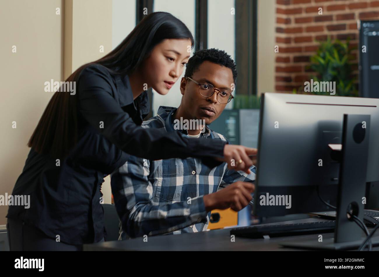 Two software developers sit at a desk with a laptop coding interface, pointing at computer screens while discussing algorithms and source code. Stock Photo