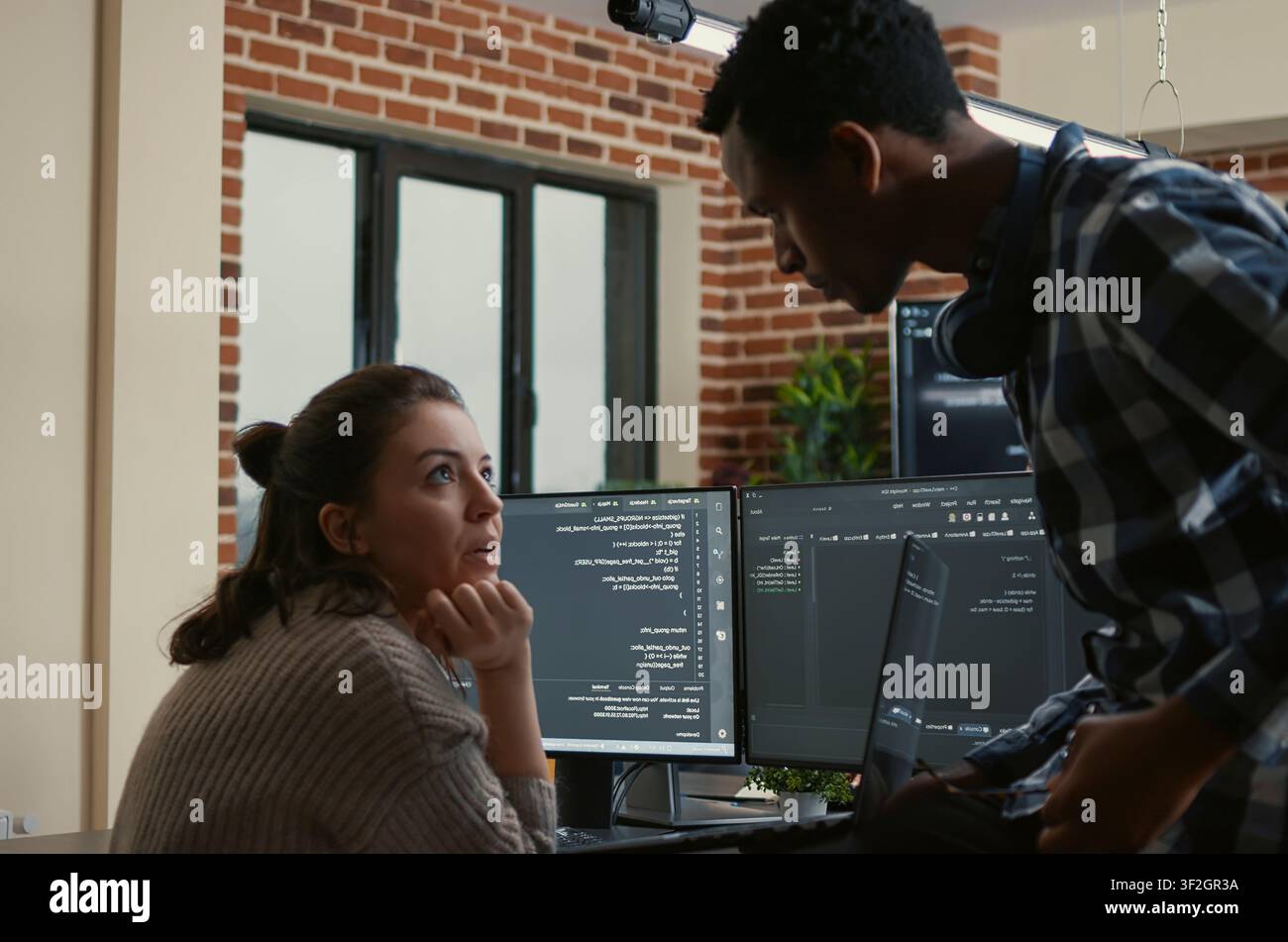 Junior developer sits on a desk and shows a laptop with source code to a senior programmer, interrupting algorithm work to ask for help fixing errors. Stock Photo