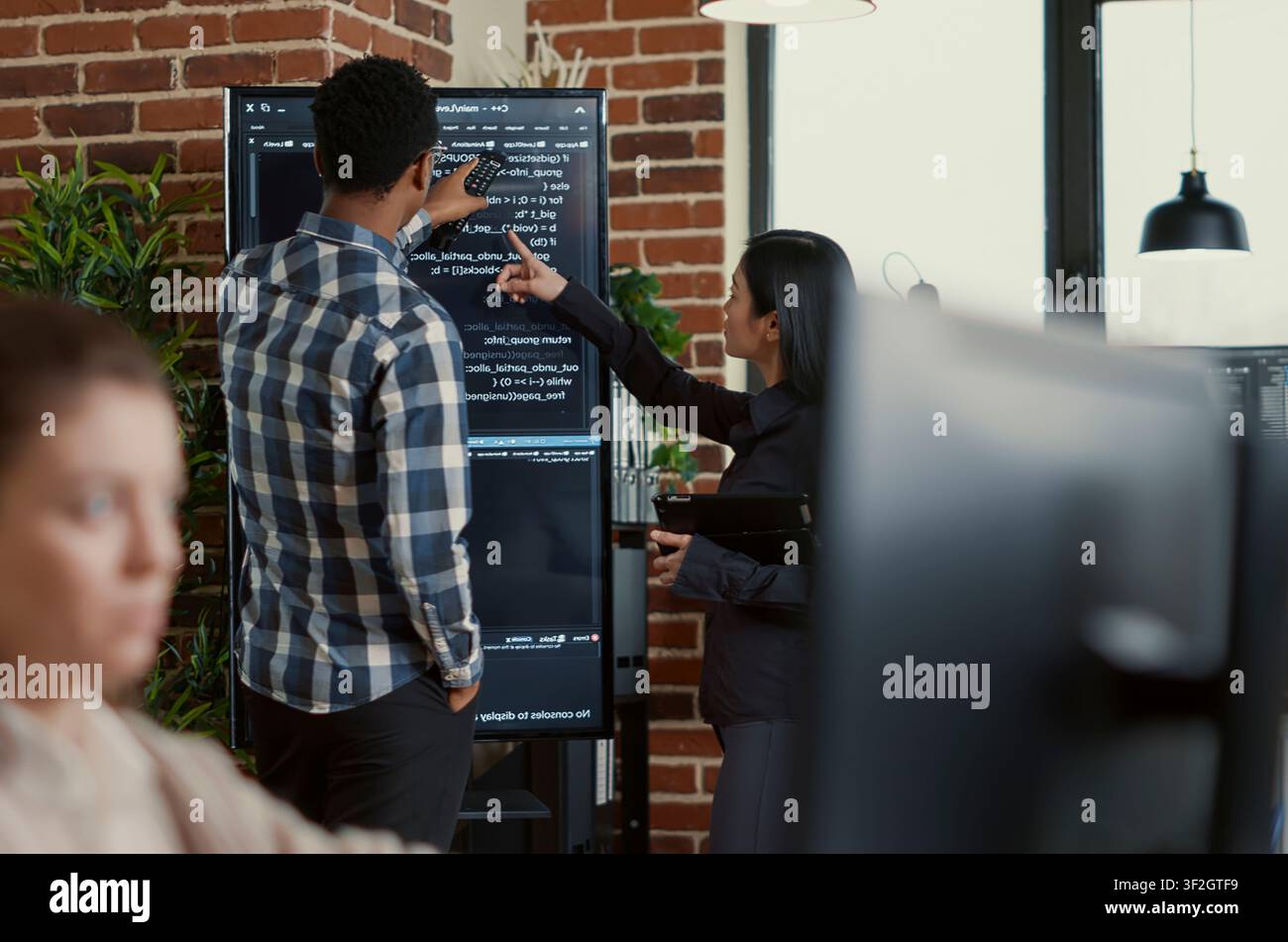 System developers analyze code on a wall screen while checking a digital tablet for errors, comparing results with a team of coders in a startup office. Stock Photo