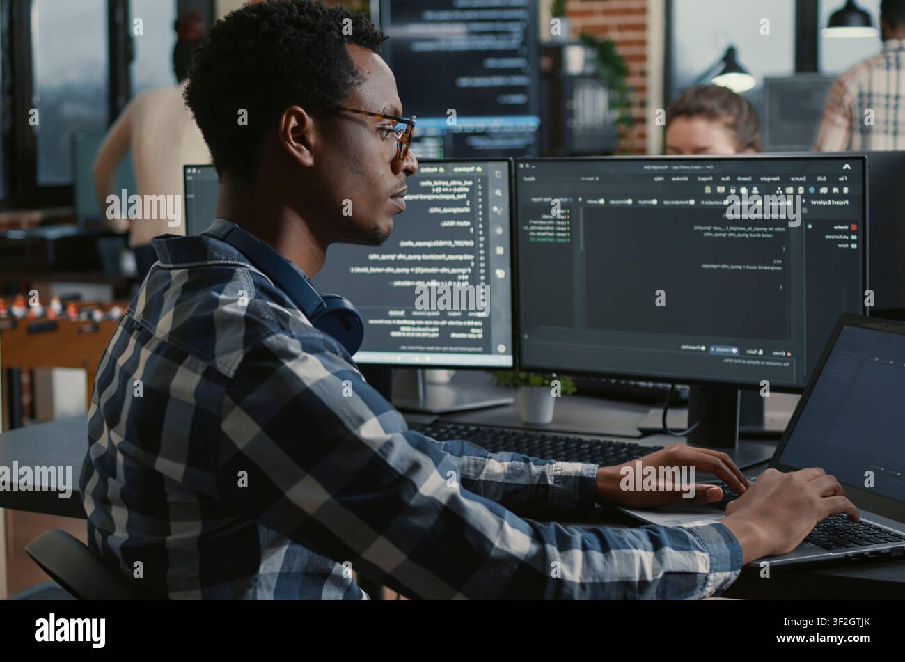 Focused programmer adjusts glasses while typing on a laptop, parsing code on multiple screens as teammates work on big data and user interface tasks in the background. Stock Photo
