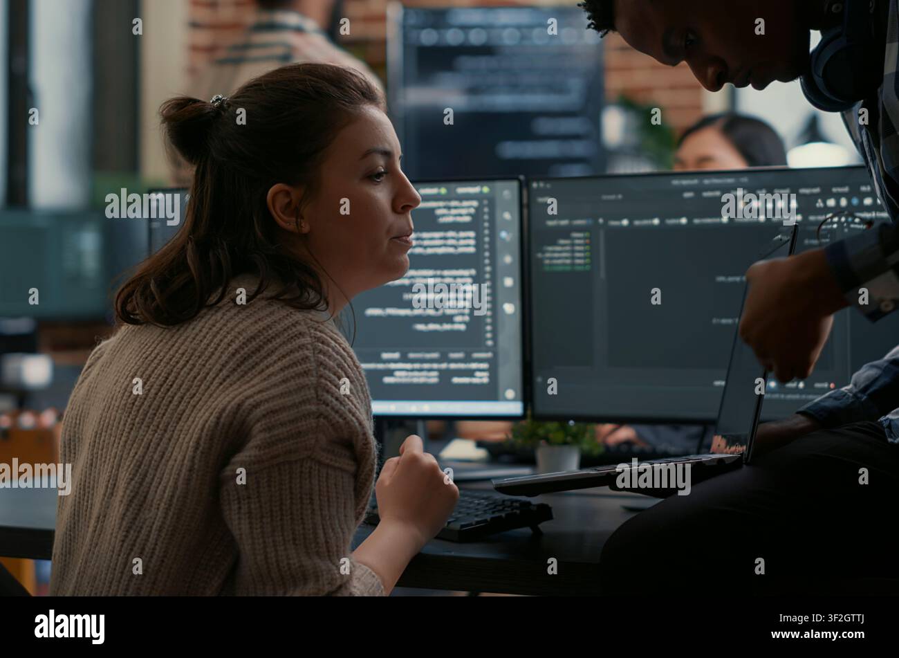 Junior programmer sits on a desk and shows laptop source code to a senior developer, asking for help with errors during algorithm work. Stock Photo