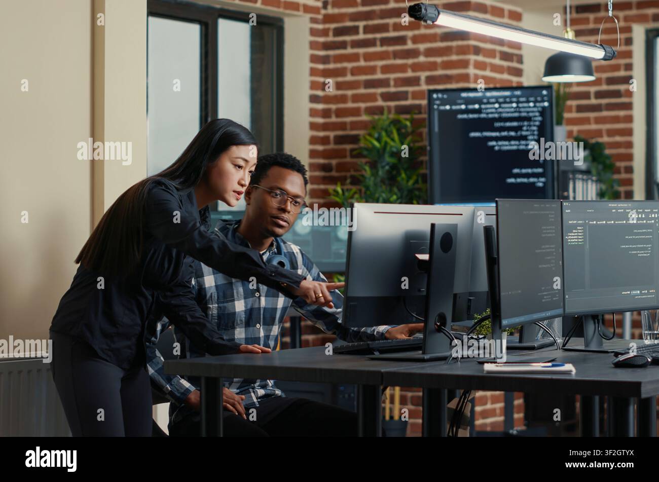 Two software developers carry a laptop with coding interface toward a desk, discussing algorithms and a group project with screens nearby. Stock Photo