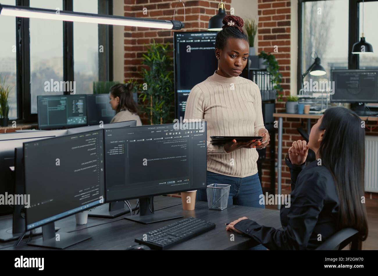 Software developer standing up holding digital tablet talking with programmer sitting at desk with computer screens showing running code around computer screens, showing developer teamwork, source. Stock Photo